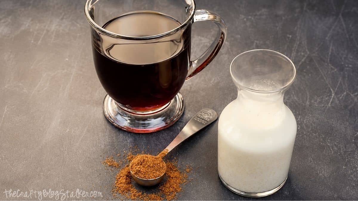 Glass mug of black coffee beside a small glass pitcher of pumpkin pie creamer and a measuring spoon of pumpkin spice on a dark countertop.