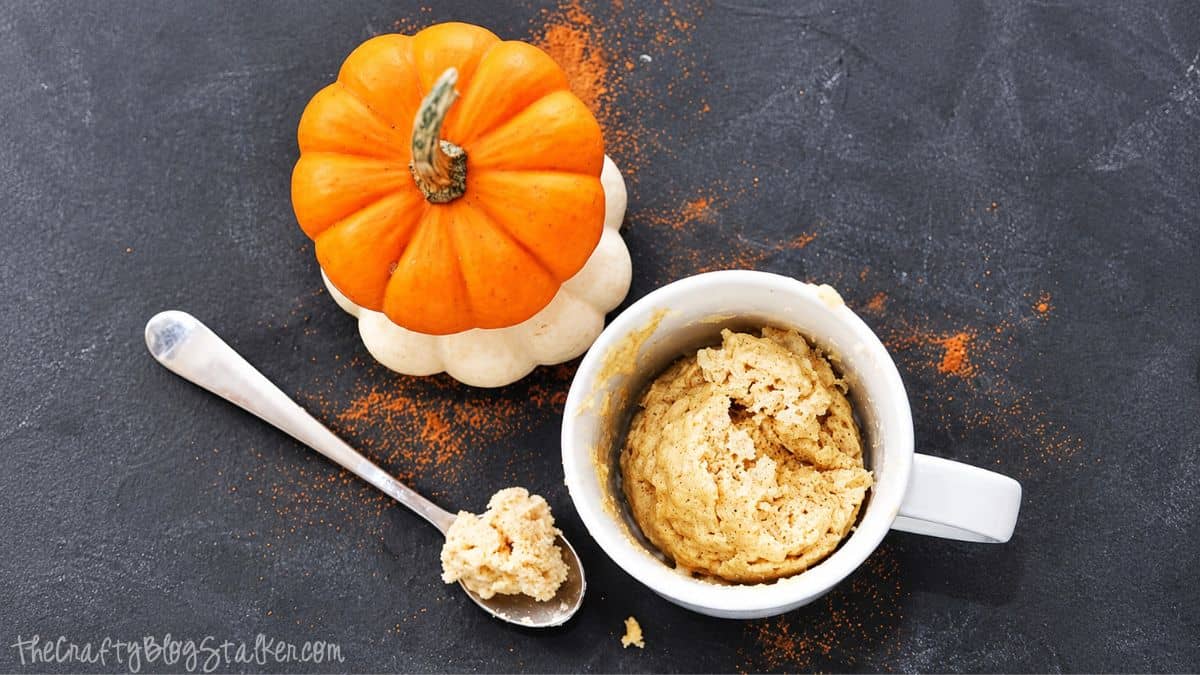 Overhead view of a pumpkin spice mug cake in a white mug with a spoonful of cake, two small pumpkins, and pumpkin spice sprinkled nearby.