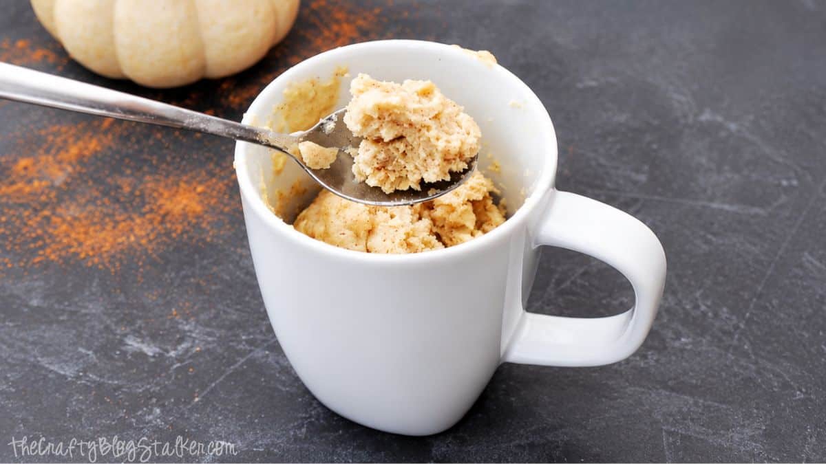 Spoonful of pumpkin spice mug cake held above a white mug with more cake inside, set on a dark surface with a small pumpkin in the background.