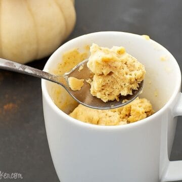 Spoonful of pumpkin spice mug cake held above a white mug with more cake inside, set on a dark surface with a small pumpkin in the background.