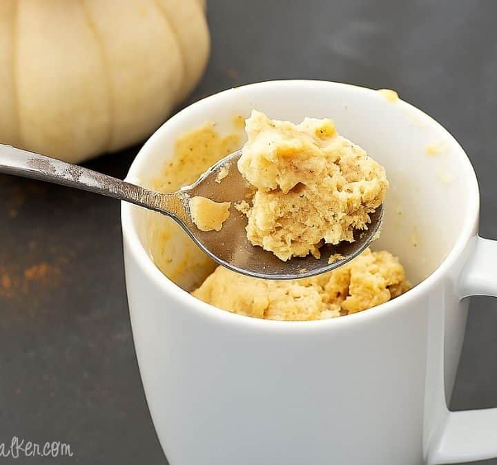 Spoonful of pumpkin spice mug cake held above a white mug with more cake inside, set on a dark surface with a small pumpkin in the background.