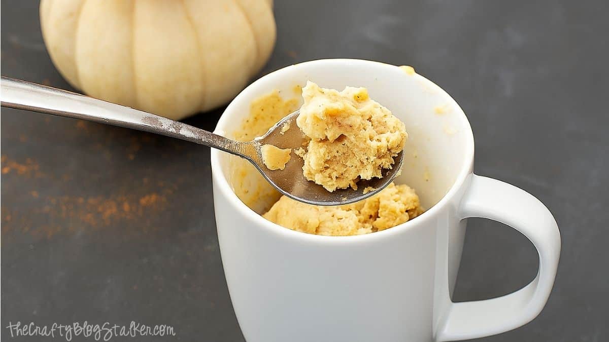 Spoonful of pumpkin spice mug cake held above a white mug with more cake inside, set on a dark surface with a small pumpkin in the background.