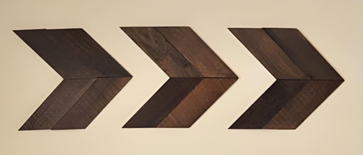 Wall art featuring three dark wood arrows arranged in a row on a light-colored background, each made of two angled wood pieces forming a chevron shape.
