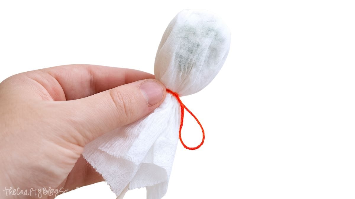 A hand holds a lollipop wrapped in white tissue, tied with orange string to form a ghost shape for a Halloween craft.