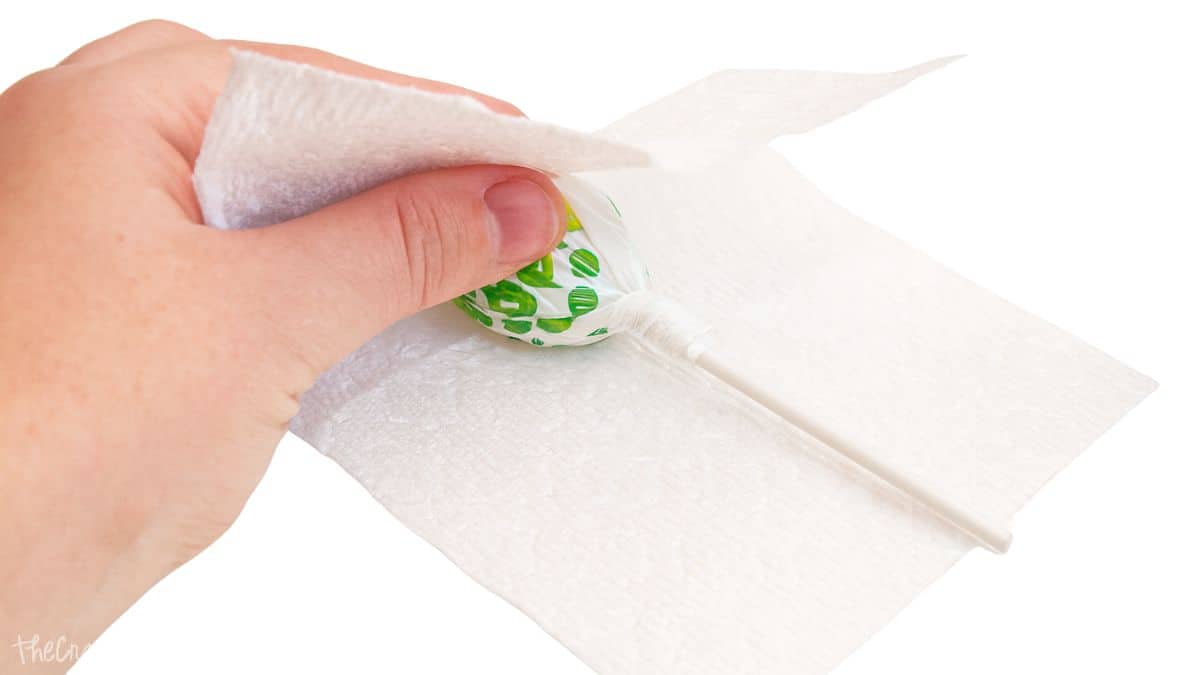 A hand wraps a green and white lollipop in a piece of white tissue paper on a white surface.