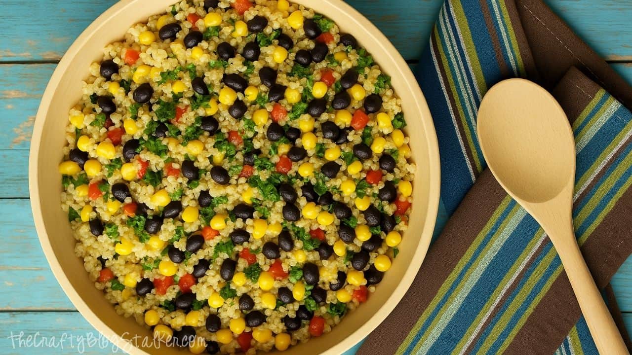 A wooden bowl filled with black bean, corn, and quinoa salad sits on a turquoise table next to a striped napkin and wooden spoon.