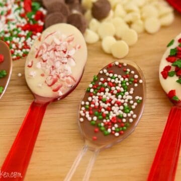 Four chocolate spoons topped with crushed candy canes and red, green, and white sprinkles, displayed on a wooden table with chocolate chips in the background.