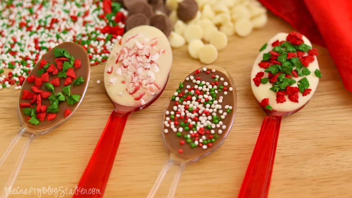 Four chocolate spoons topped with crushed candy canes and red, green, and white sprinkles, displayed on a wooden table with chocolate chips in the background.