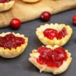 Mini phyllo tartlets filled with melted brie and cranberry sauce on a dark surface, with whole cranberries scattered around.