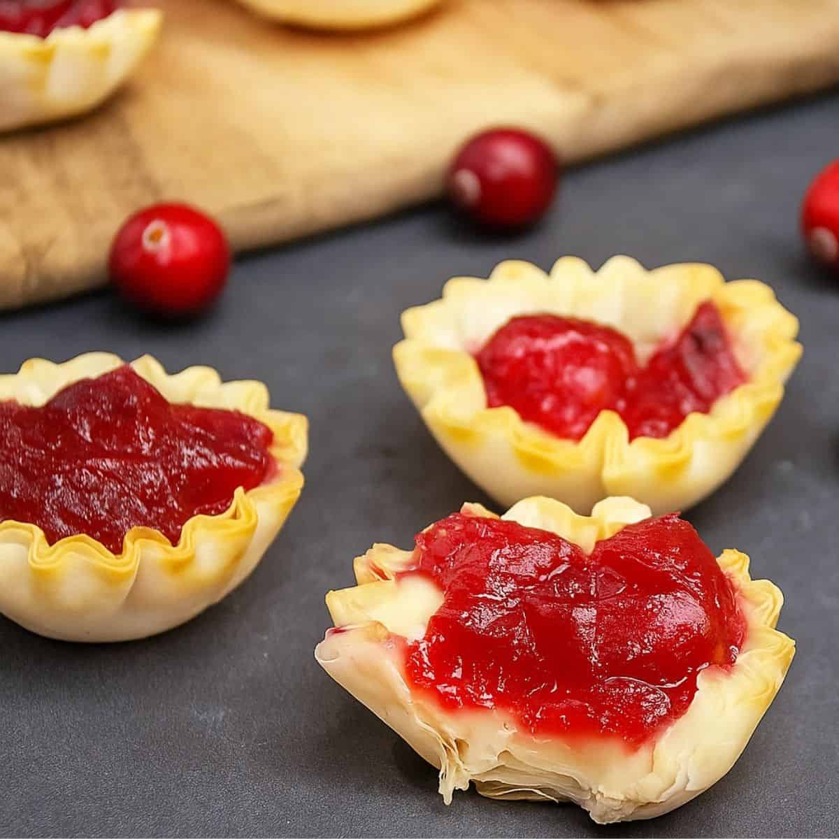 Mini phyllo tartlets filled with melted brie and cranberry sauce on a dark surface, with whole cranberries scattered around.