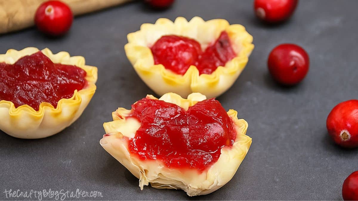 Mini phyllo tartlets filled with melted brie and cranberry sauce on a dark surface, with fresh cranberries scattered around.