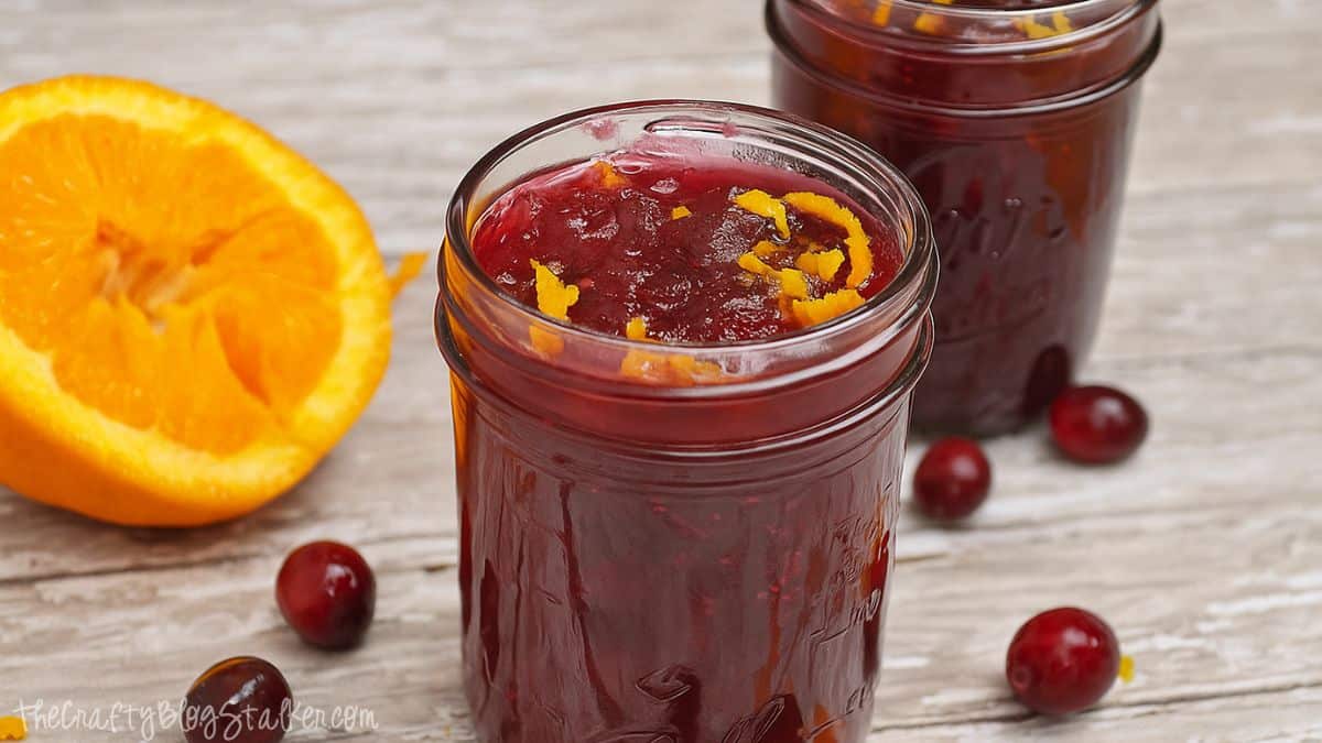 Jar of homemade cranberry sauce topped with orange zest, surrounded by fresh cranberries and a sliced orange on a rustic wooden surface.