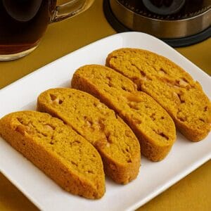 Pumpkin biscotti slices arranged on a white plate, with a warm drink partially visible in the background.