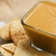 Square glass bowl filled with creamy pumpkin pie dip surrounded by cinnamon pita chips on a clear plate set over a burlap surface.