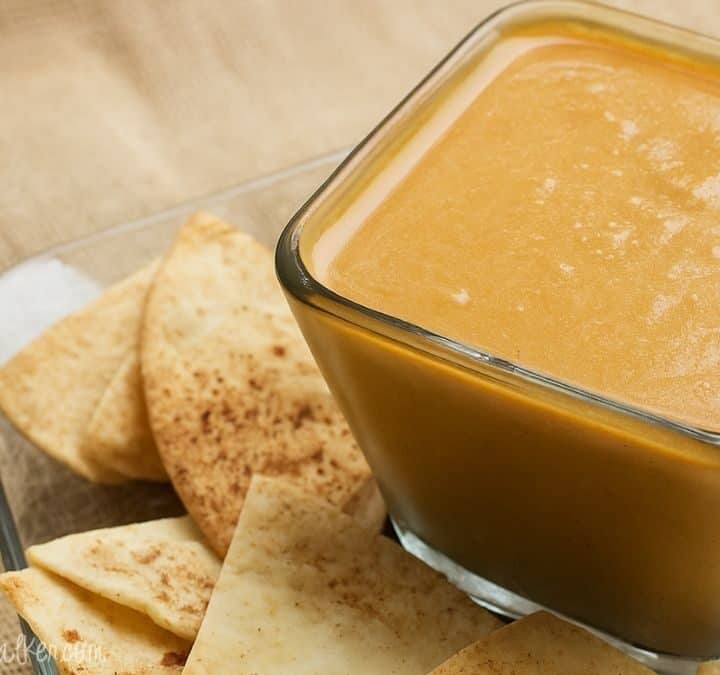 Square glass bowl filled with creamy pumpkin pie dip surrounded by cinnamon pita chips on a clear plate set over a burlap surface.