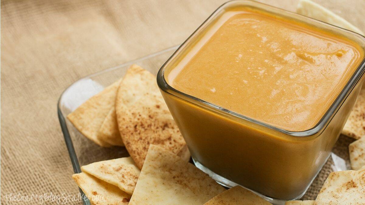 Square glass bowl filled with creamy pumpkin pie dip surrounded by cinnamon pita chips on a clear plate set over a burlap surface.