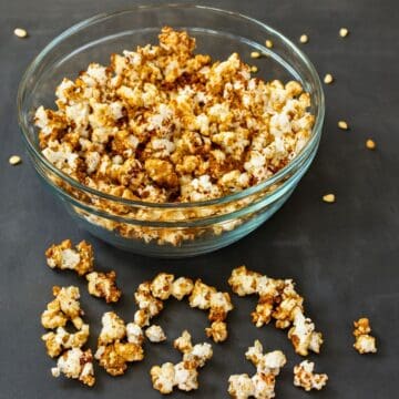 Glass bowl filled with pumpkin pie popcorn, with caramel-coated popcorn pieces scattered on a dark surface beside the bowl.