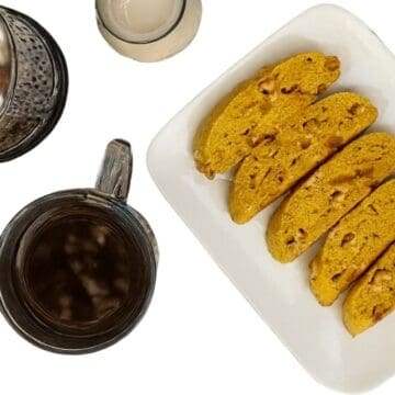 Plate of white chocolate pumpkin biscotti beside a cup of coffee, a glass of creamer, and a French press on a white background.