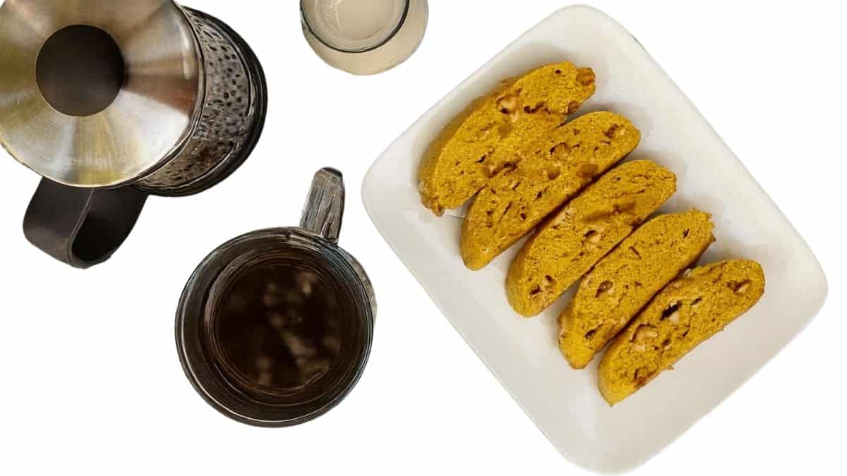 Plate of white chocolate pumpkin biscotti beside a cup of coffee, a glass of creamer, and a French press on a white background.