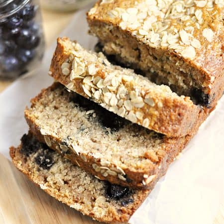 Sliced blueberry oatmeal bread topped with oats, showcasing its moist texture and juicy blueberries, placed on parchment paper with fresh blueberries in the background.