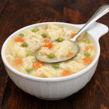 Bowl of homemade chicken and dumplings with tender chicken, fluffy dumplings, peas, and carrots in a creamy broth, served with a spoon on a wooden table.