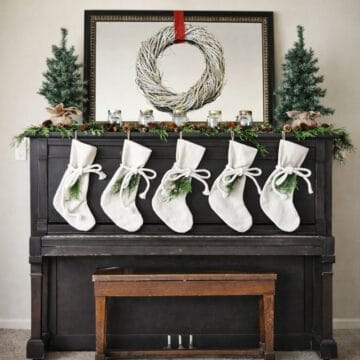 Upright piano decorated for Christmas with a wreath, greenery garland, small trees, candles, and five stockings hanging across the front.