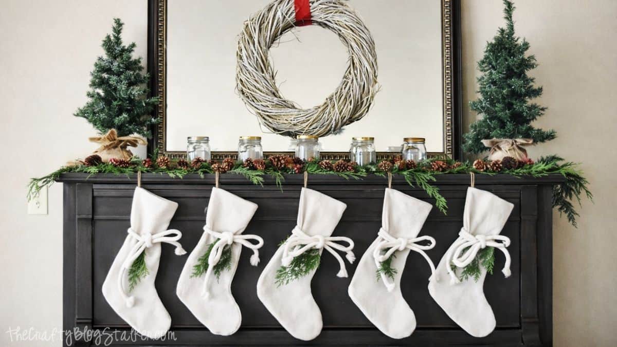 A Rustic Christmas Mantel featuring real cedar garland and drop cloth stockings Upright piano decorated with a wreath, greenery garland, mason jars, small trees, and five stockings hanging across the front for Christmas.