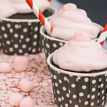 Chocolate cupcakes topped with fluffy pink peppermint marshmallow frosting, served in black polka dot cups with red and white striped straws.