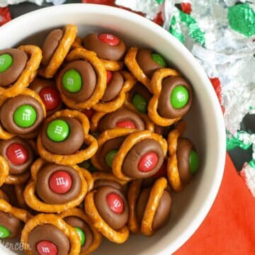 Bowl of holiday pretzel bites made with pretzels, melted chocolate kisses, and red and green M&M candies, surrounded by festive foil wrappers.