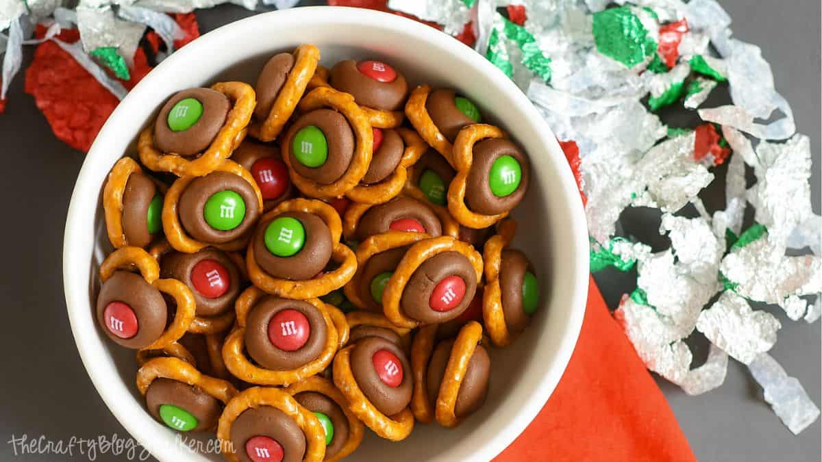 Bowl of holiday pretzel bites made with pretzels, melted chocolate kisses, and red and green M&M candies, surrounded by festive foil wrappers.