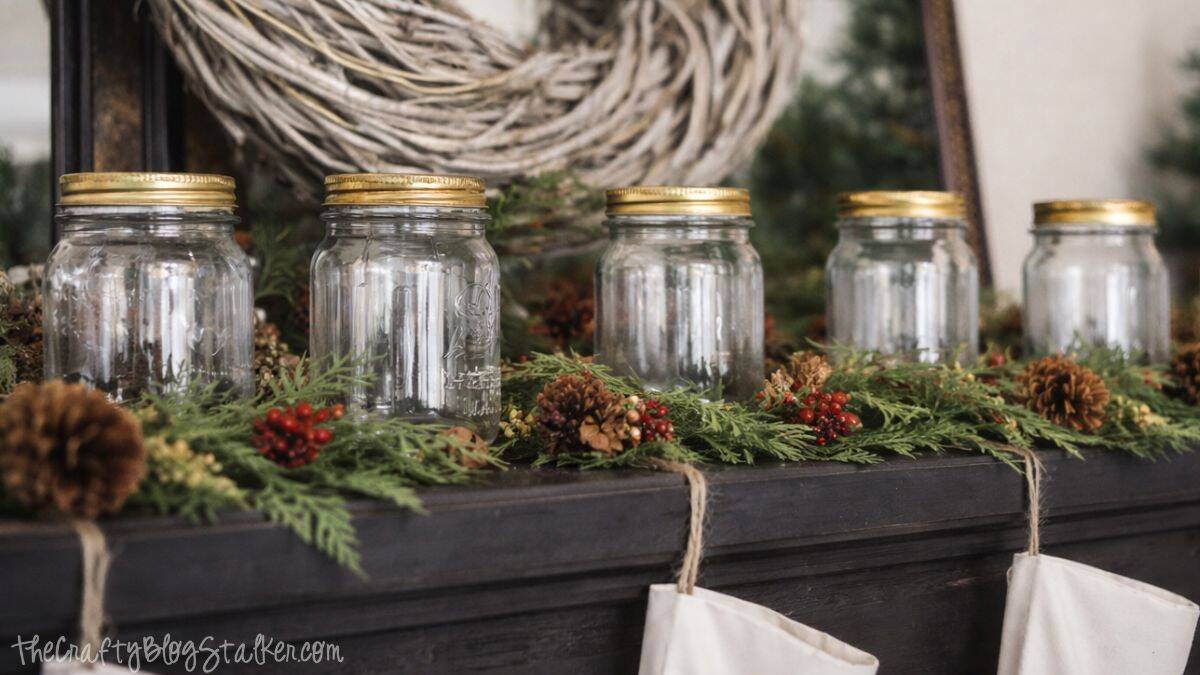 A Rustic Christmas Mantel featuring real cedar garland and drop cloth stockings Mason jars lined up on a piano mantel with cedar greenery, pinecones, and berry accents as part of a rustic Christmas decor setup.