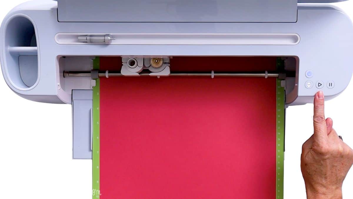 Valentine Treat Bag Tutorial #Valentine #PaperCrafts A Cricut cutting machine loaded with a cardstock sheet on a cutting mat, with a hand pressing the start button to begin cutting.