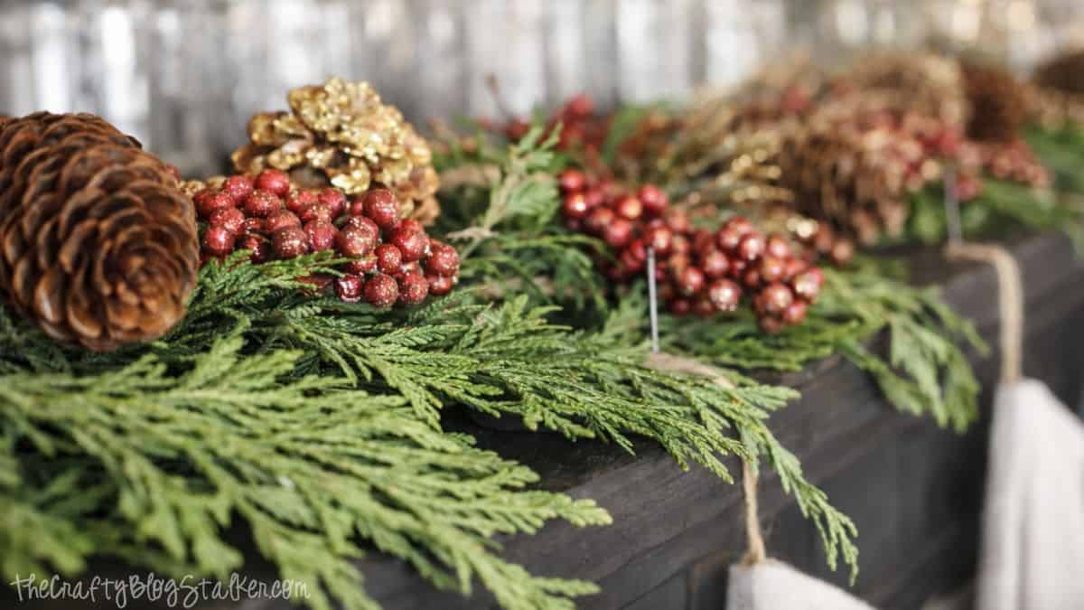 A Rustic Christmas Mantel featuring real cedar garland and drop cloth stockings Close-up of cedar garland with pinecones and berry accents arranged along the top of a piano as part of Christmas mantel decor.