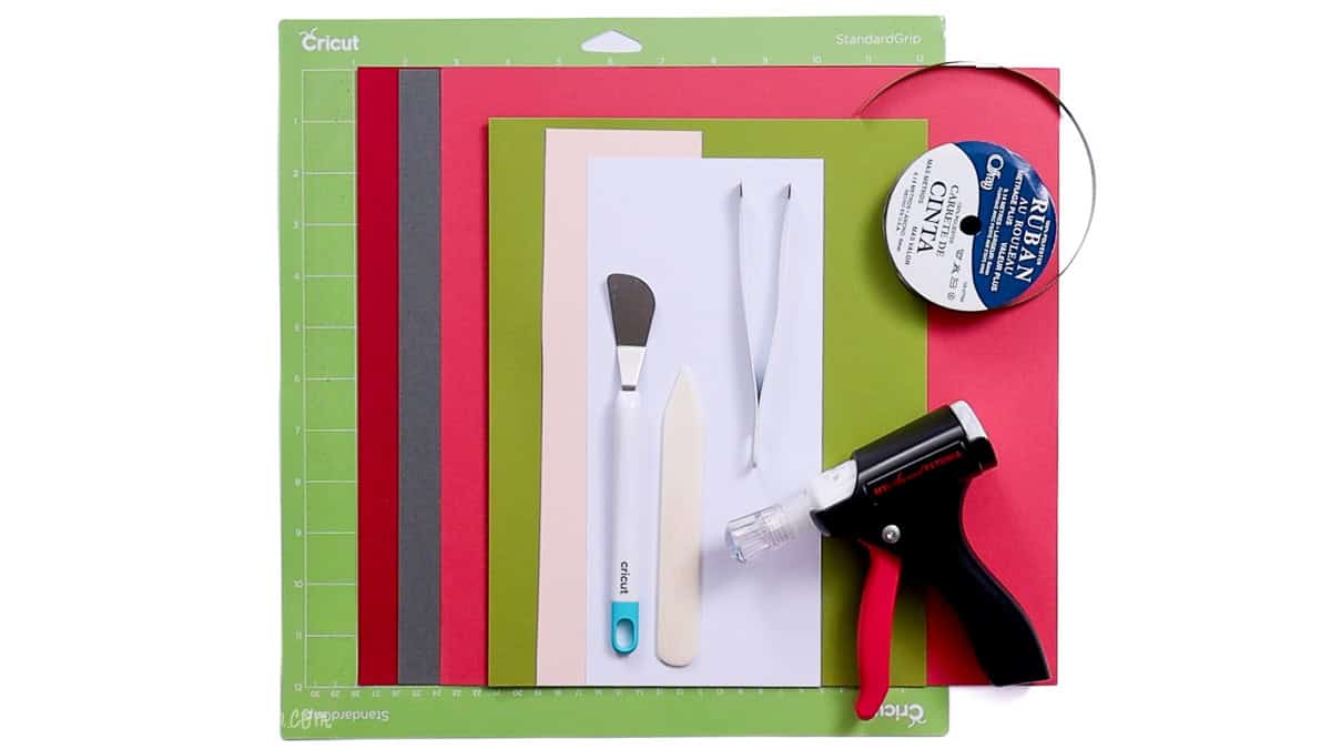 Supplies for making Valentine treat bags including cardstock sheets, a Cricut cutting mat, ribbon, glue gun, weeding tools, and crafting spatulas.