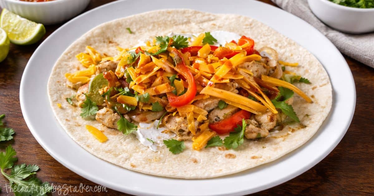 Chicken fajita tortilla topped with sliced peppers, onions, shredded cheese, cilantro, and sour cream on a white plate with lime wedges nearby.