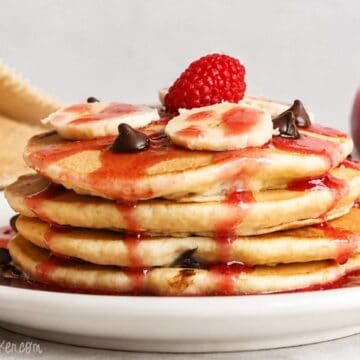 Stack of chocolate chip pancakes topped with banana slices, chocolate chips, raspberry syrup, and a fresh raspberry on a white plate.