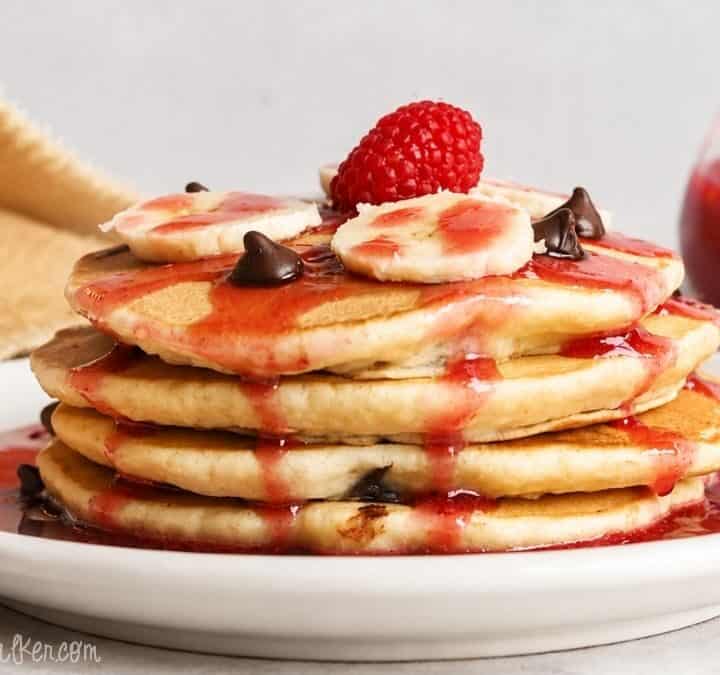 Stack of chocolate chip pancakes topped with banana slices, chocolate chips, raspberry syrup, and a fresh raspberry on a white plate.