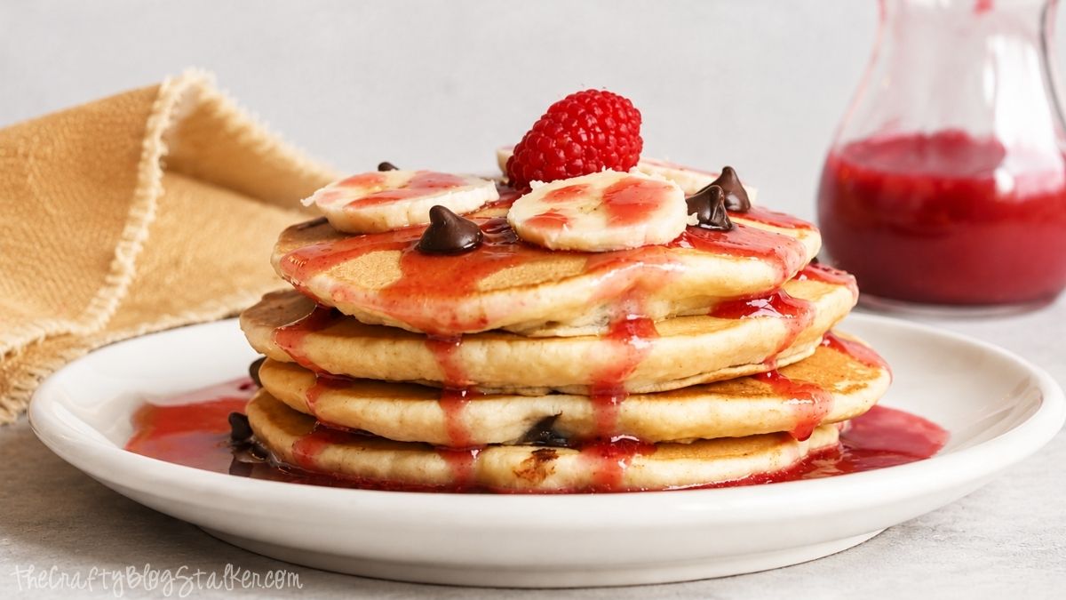 Stack of chocolate chip pancakes topped with banana slices, chocolate chips, raspberry syrup, and a fresh raspberry on a white plate.