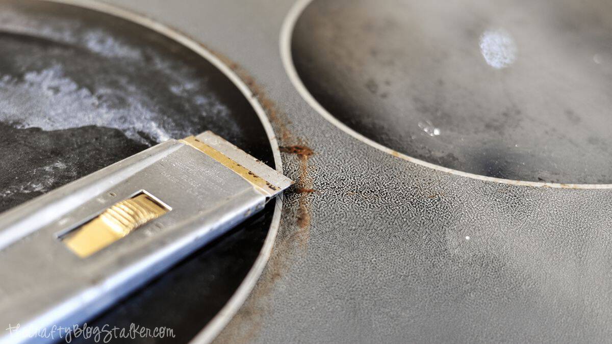 Close-up of a scraper removing burned-on residue from a dirty glass top stove during the cleaning process.