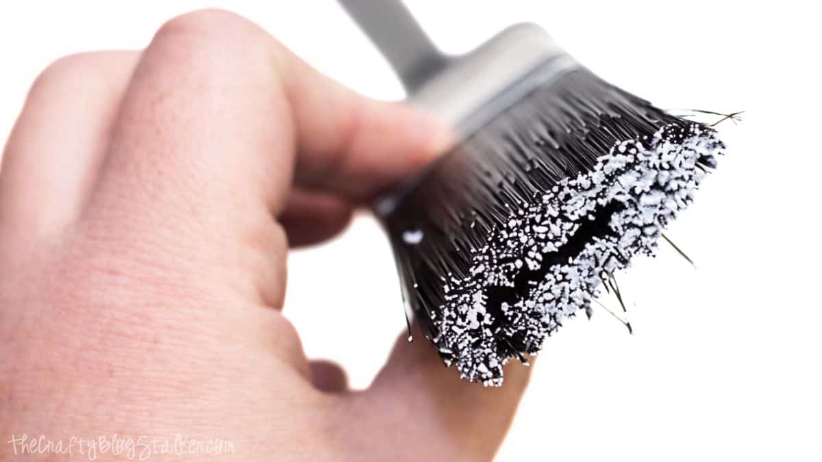 Close-up of a hand holding a paintbrush with dry brush technique, showing lightly loaded bristles coated with white paint.