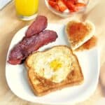 A breakfast plate with heart-shaped egg-in-toast, crispy bacon, and heart-cut toast with jam, served with orange juice and fresh strawberries in a bowl.