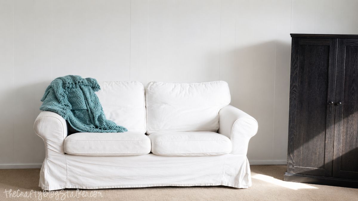 White slipcovered loveseat with a teal throw blanket sits against freshly painted white wood paneling beside a dark wood cabinet.