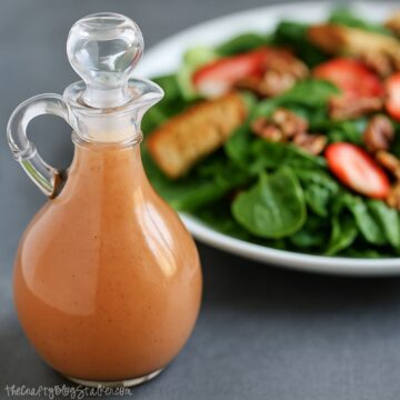 Glass bottle of homemade strawberry vinaigrette with a fresh spinach salad topped with strawberries, pecans, and croutons in the background.