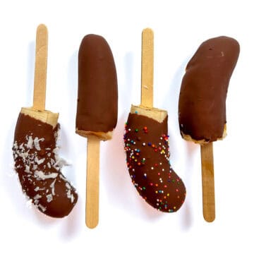 Four frozen banana popsicles dipped in chocolate, topped with shredded coconut and colorful sprinkles, arranged on wooden sticks against a white background.