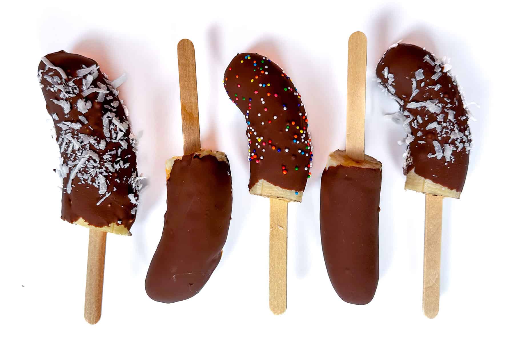 Five frozen banana popsicles dipped in chocolate, topped with shredded coconut and colorful sprinkles, displayed on wooden sticks against a white background.