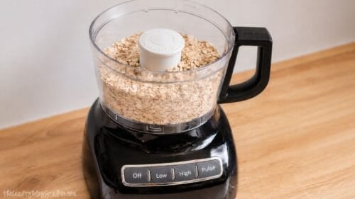 A black food processor filled with rolled oats on a wooden countertop, ready to blend into homemade oat flour.