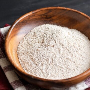 A wooden bowl filled with homemade oat flour sits on a red and white checkered kitchen towel on a dark wooden table.