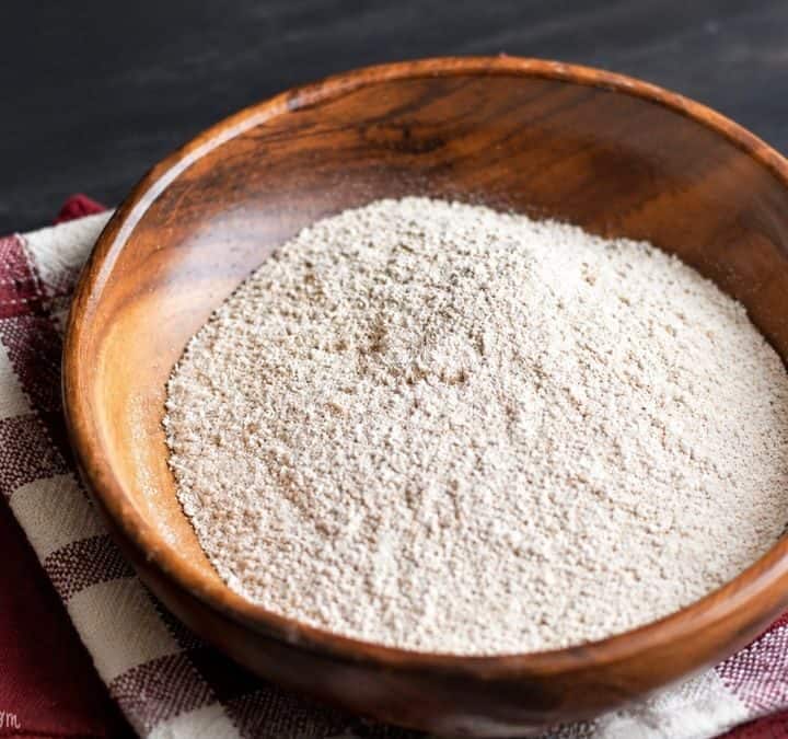 A wooden bowl filled with homemade oat flour sits on a red and white checkered kitchen towel on a dark wooden table.