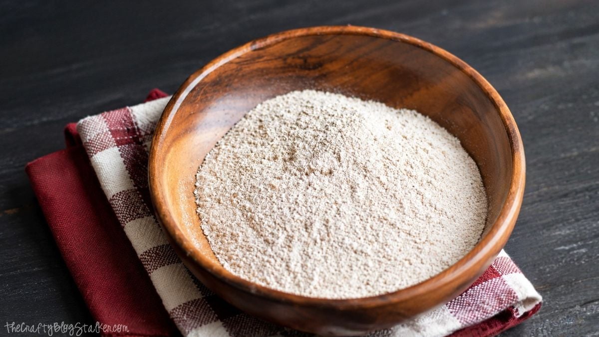A wooden bowl filled with homemade oat flour sits on a red and white checkered kitchen towel on a dark wooden table.