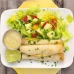 Plate with oven-baked chicken taquitos, fresh lettuce salad topped with avocado and tomatoes, lime wedges, and creamy dipping sauce.